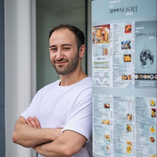Smiling dark haired man wearing a white t shirt leaning against doorframe beside menu board for Moonlight Cafe