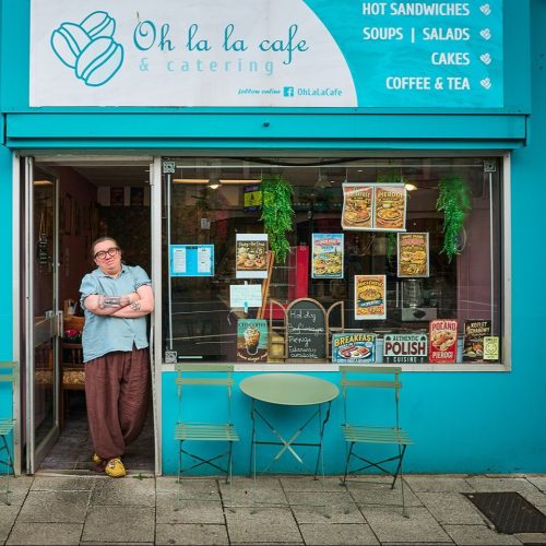 Blue cafe front with smiling lady in doorway, with her arms crossed, signage says Oh La La Cafe, table and chairs outside.