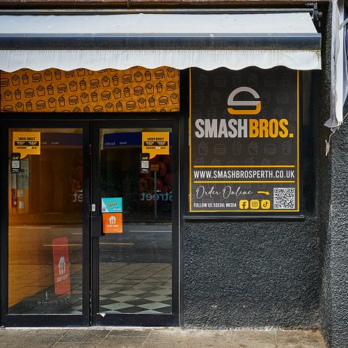A black building with a yellow board with burger and fries decals over it above the door. There is a large sign which says 'Smash Bros.'