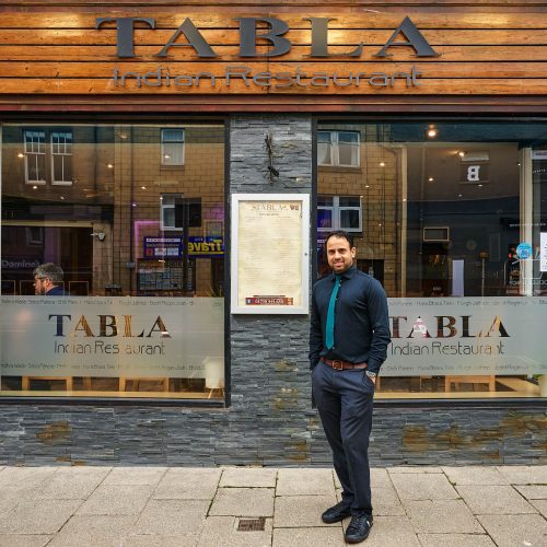 A smiling man wearing a navy shirt with a teal tie standing outside a grey brick building with a wooden sign board which reads 'Tabla'