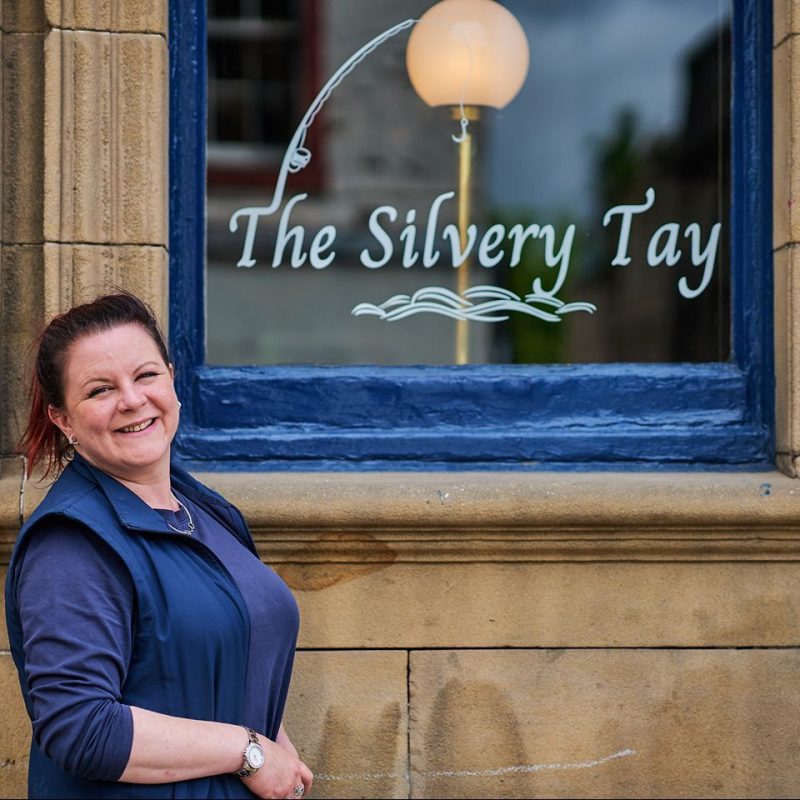 Smiling lady with dark hair beside window that has The Silvery Tay written on it.