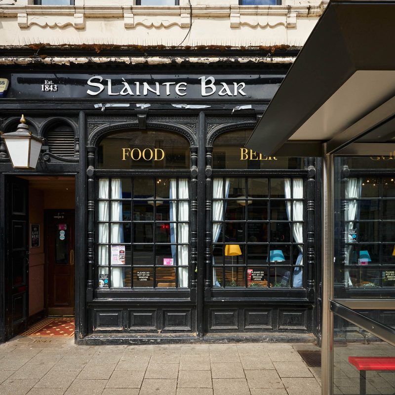 A black building with square windows and a white sign which says 'Slainte Bar'