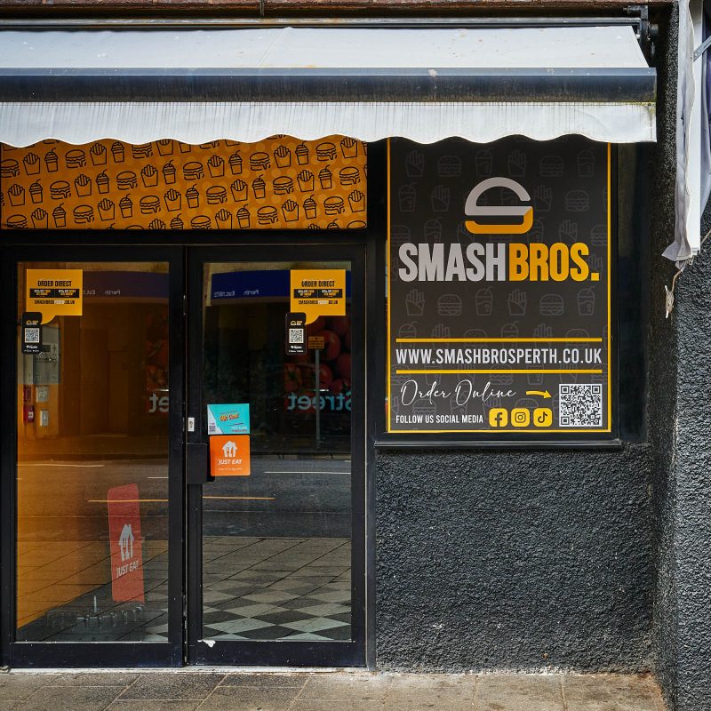 A black building with a yellow board with burger and fries decals over it above the door. There is a large sign which says 'Smash Bros.'