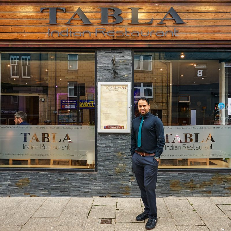 A smiling man wearing a navy shirt with a teal tie standing outside a grey brick building with a wooden sign board which reads 'Tabla'