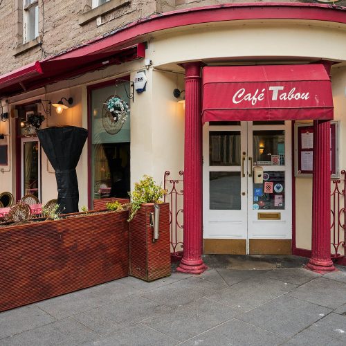 A white building with red accents and collumns. The sign says Cafe Tabou. There are brown wooden plant boxes with flowers in them.