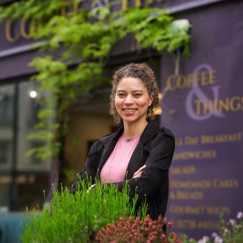 A smiling women wearing a pink t-shirt and balck blazer standing outside a purple building with the words Coffee & Things painted on it in lilac.
