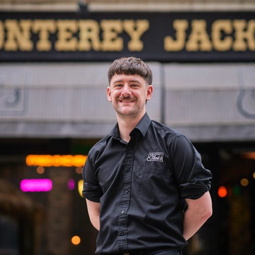 A smiling man wearing a black shirt which has 'Monterery Jack's' embrodiered on it in white lettes. He is standing in front of a building which has a black and gold sign which also reads 'Monterery Jack's