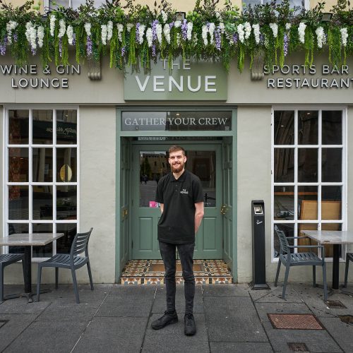 A smiling man wearing a black polo shirt which reads 'The Venue' in white embroidery is standing in front of a beige building with sage coloured accents