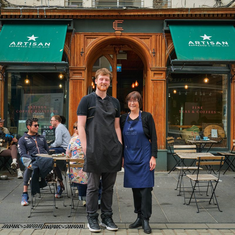 A smiling man in a black polo shirt and black apron standing next to a smiling woman wearing a black long sleeved t-shirt and a navy blue apron with the words Artisan Espresso & Wine Bar on it.