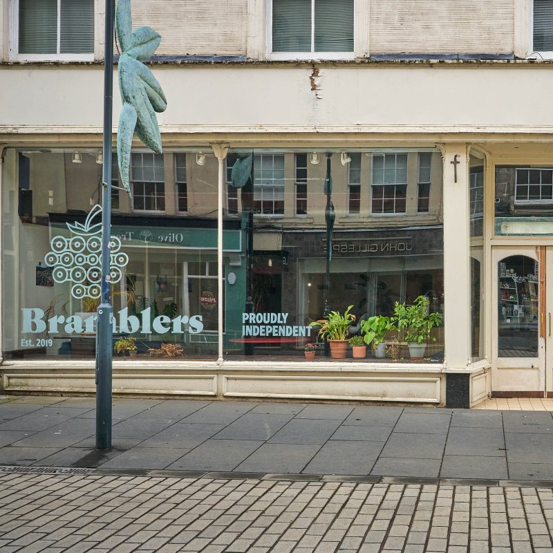 A beige building with big tall windows. There are decals on the windows which say 'Bramblers. Proudly Independent' Above this there is a decal of a blackberry.