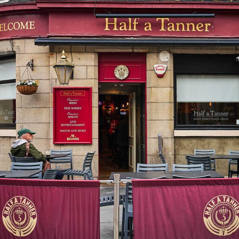 A brick building with Red accents and a gold sign which reads 'Half a Tanner.' There are outdoor grey seating where a man in a green cap and jacket is sitting.