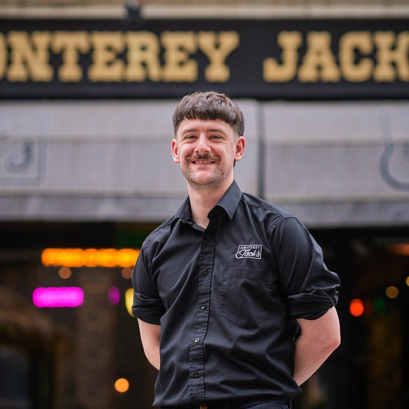 A smiling man wearing a black shirt which has 'Monterery Jack's' embrodiered on it in white lettes. He is standing in front of a building which has a black and gold sign which also reads 'Monterery Jack's