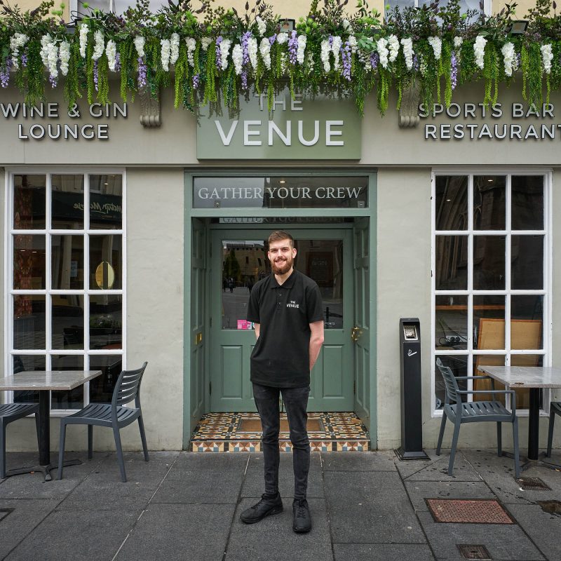 A smiling man wearing a black polo shirt which reads 'The Venue' in white embroidery is standing in front of a beige building with sage coloured accents