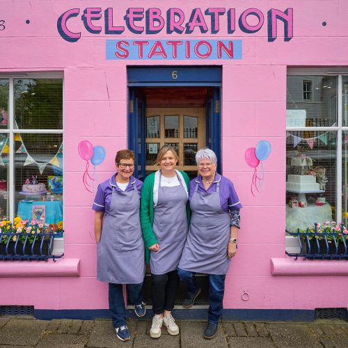 Three smiling women wearing purple polo shirts standing on front of a hot pink building. There are balloons painted on the side.