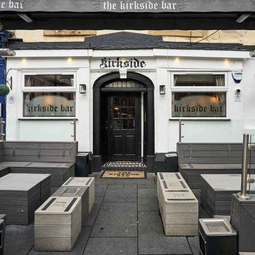 A white building with a black doorway, It has some dark grey outdoor seating.