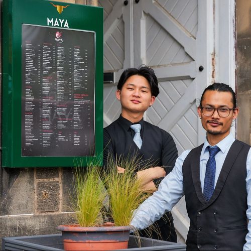 two smiling men wearing waistcoats and shirts and ties beside menu board for Mayas Japanese restaurant