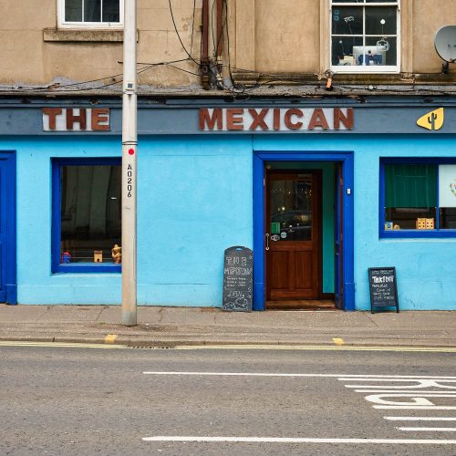 A light blue building with dark blue accents. It has a dark wooden sign which says 'The Mexican' on it.