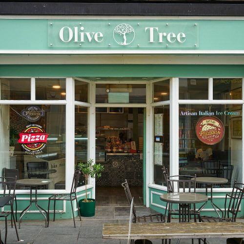 A green building with white accents. It has a white sign which reads 'Olive Tree'. In the window it has decals advertising their pizza. It also has dark grey outdoor seating.