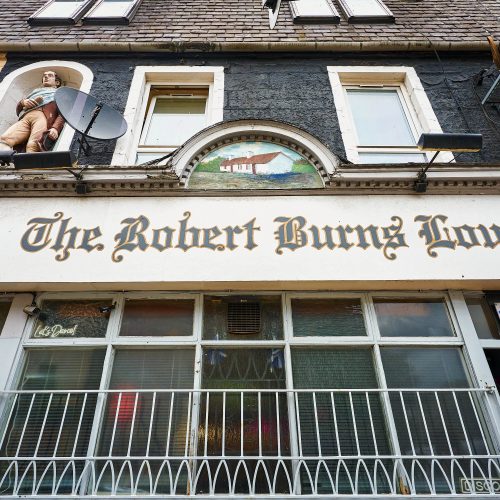 A white sign which has 'The Robert Burns Lounge' written in black and gold text. Above is a painted image of two cottages. There is also a statue of Robert Burns.