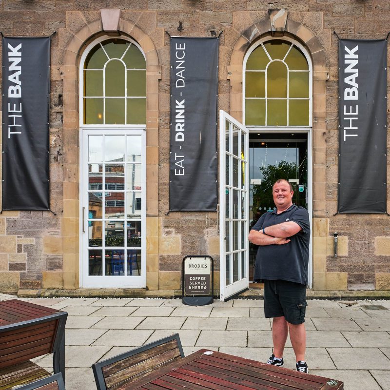 A smiling man in a black polo shirt and black shorts standing outside the Bar he has his arms crossed.