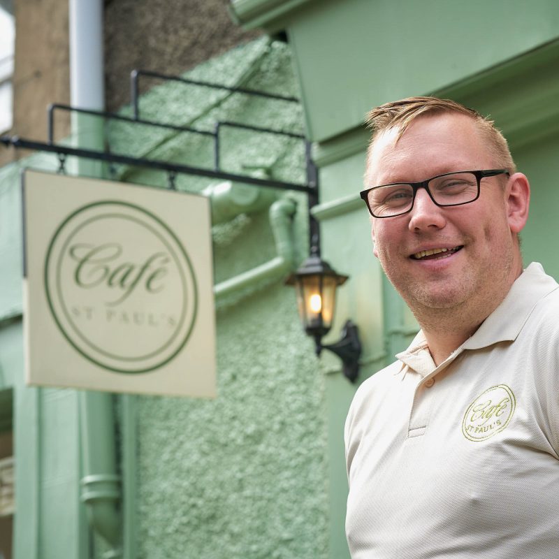 A smiling man wearing a white polo shirt which has Cafe St Paul's embroidered in gold. He is standing next to a mint coloured building and a white sign with the same gold text on it.