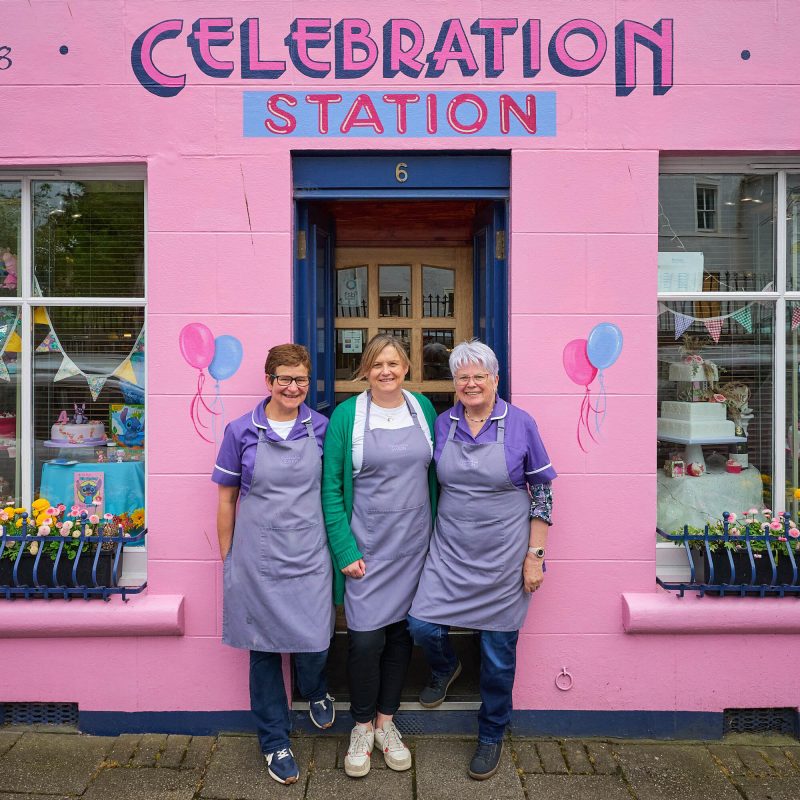 Three smiling women wearing purple polo shirts standing on front of a hot pink building. There are balloons painted on the side.