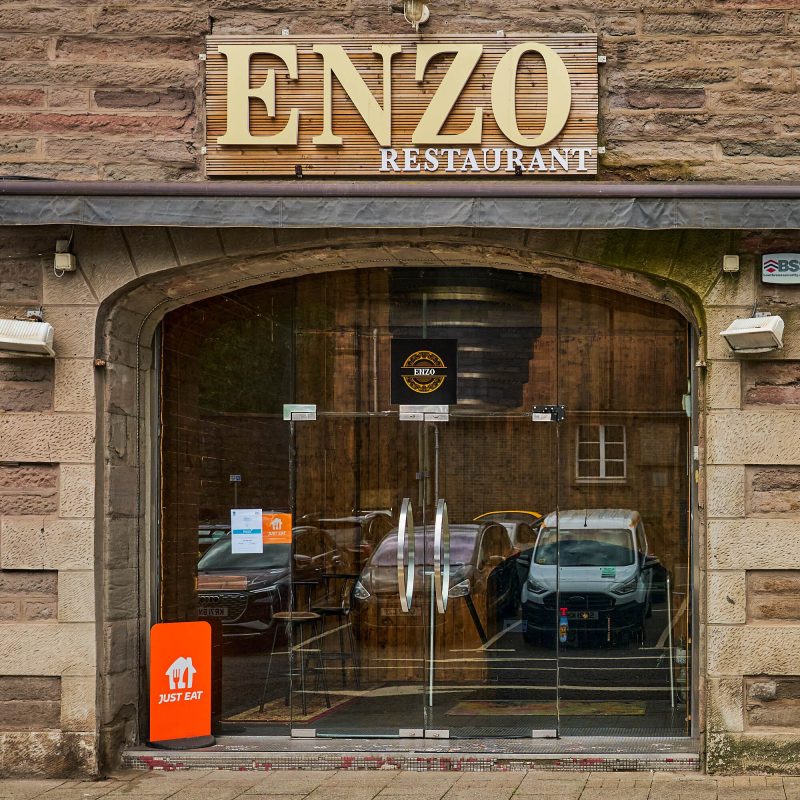 A brick building with a gold and white sign which says 'Enzo Restaurant'. It has a large glass entranceway with glass doors.