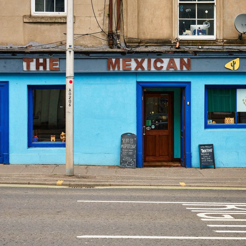 A light blue building with dark blue accents. It has a dark wooden sign which says 'The Mexican' on it.