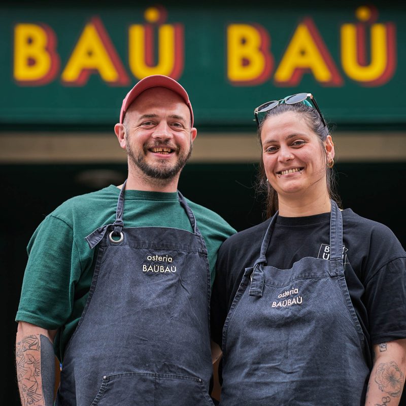 A smiling man and woman wearing denim aprons which have 'Osteria Bau Bau' embroidered on them in beige lettering. They are standing in front of a building with a green sign which reads 'Osteria Bau Bau in yellow writting.