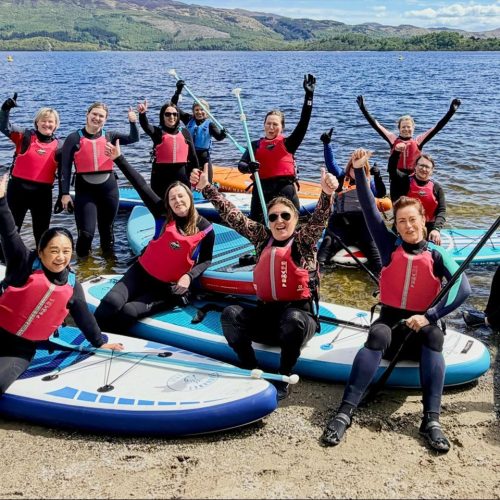 A group of smiling people wearing wetsuits and Life vests sitting atop paddleboards on a sunny day in front of a loch. Green hills in background