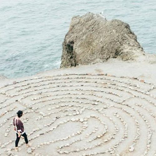 Woman walking a maze made with stones at a beach with a large rock and the sea.