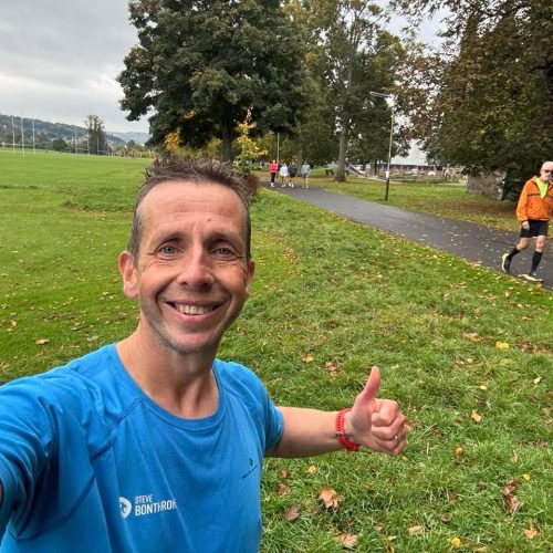 Smiling man taking a selfie with people running behind on a path in a grassy parkland, with trees.