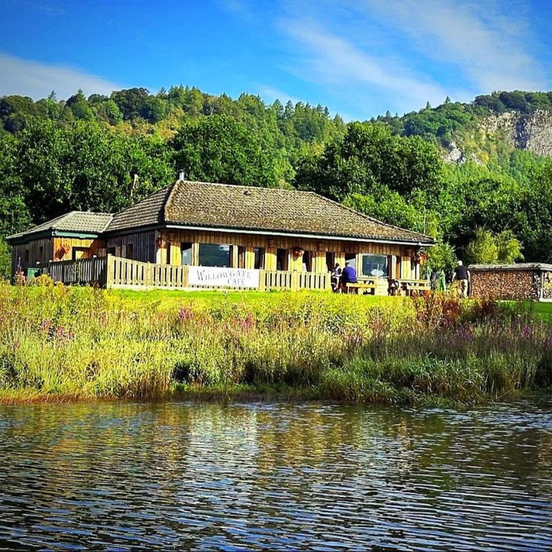 View of wooden framed single storey building on edge of River Tay, sign of fence for Willowgate Cafe, trees and hills in background on a sunny day with blue skies.
