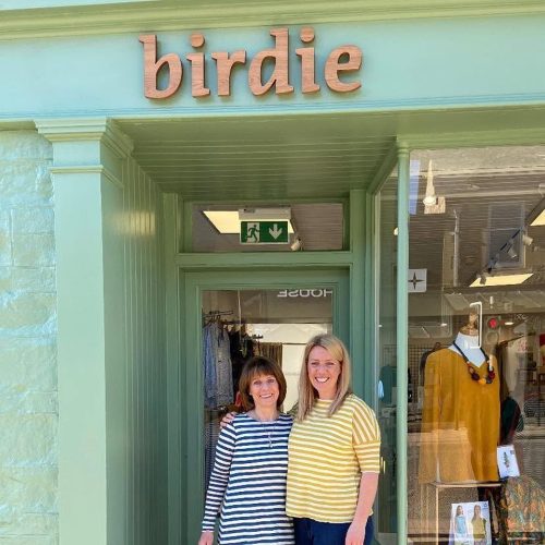 Two smiling woman wearing striped tops outside a pale green shop, underneath a sign that says Birdie