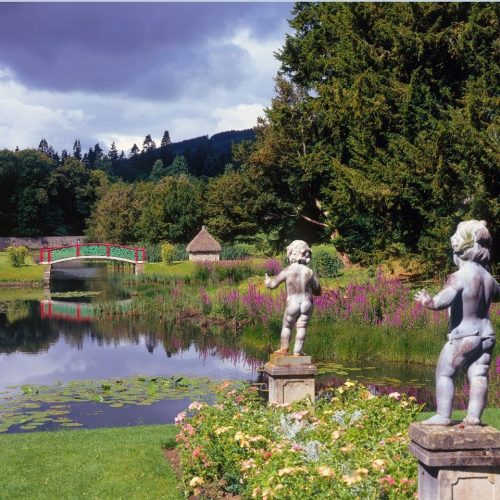Two stone cupid sculptures overlooking a large pond with a red and green decorative bridge, surrounded by bright flowers and lush green trees.