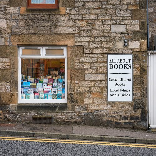 A brick building with a white sign which reads 'All About Books'. There is also a window which has secondhand books in it.