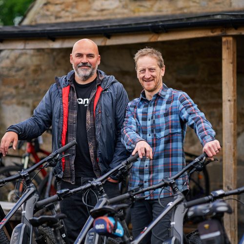 Two smiling men standing next to some bikes.