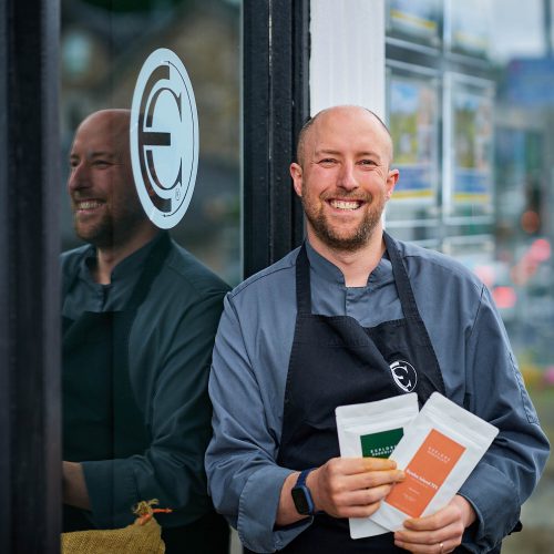 A smiling man wearing an apron with the explore chocolate logo on it. He is also holding different packets of chocolate.