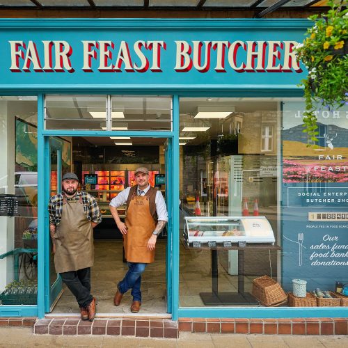 Two smiling men wearing aprons which say 'Fair Feast' on them standing in front of the Butchers shop.