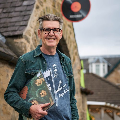 A smiling man holding two records standing outside of a brick building.