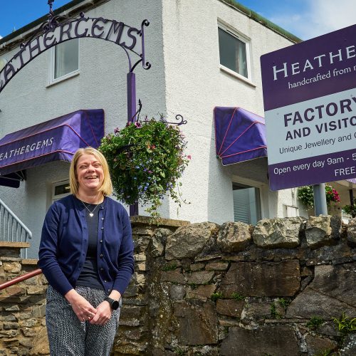 A smiling woman standing in front of a stone wall and archway which says 'Heathergems' across it.