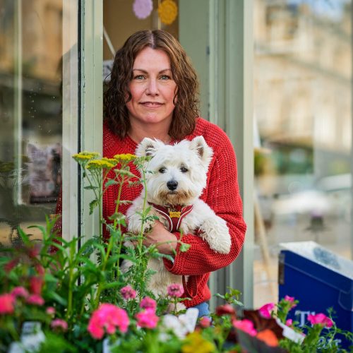 A smiling woman holding a Westie dog in front of a pale green shop.
