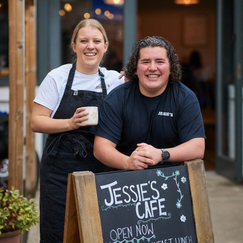 Two smiling people standing in front of a blackboard which reads Jessie's Cafe.