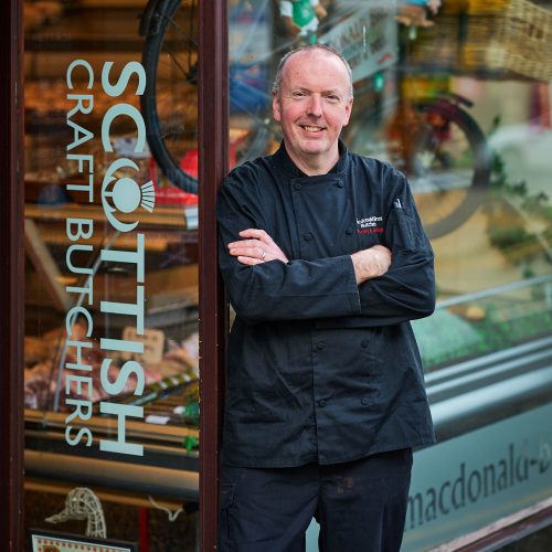 A smiling man wearing a black chefs jacket standing in front of the butchers shop.