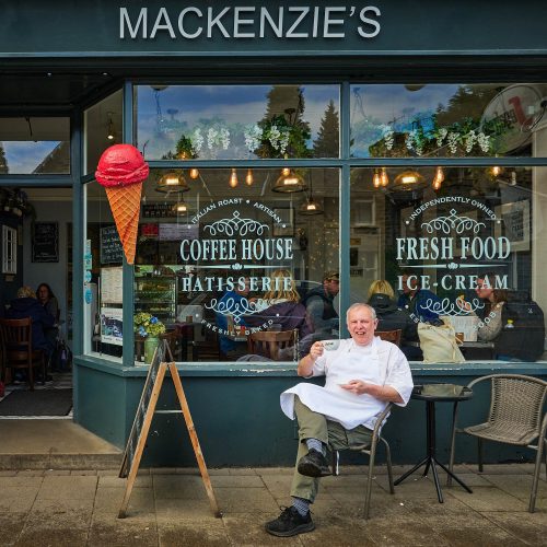 A smiling man holding a white coffee cup sitting in an outdoor chair outside the coffee shop.
