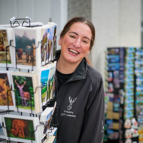 A smiling woman wearing a fleece which says 'Pitlochry Gift Company' on it. She is standing behind a postcard stand.