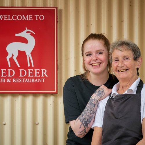 Two smiling women standing in front of a red sign which has a white deer on it and reads 'Red Deer Restaurant'