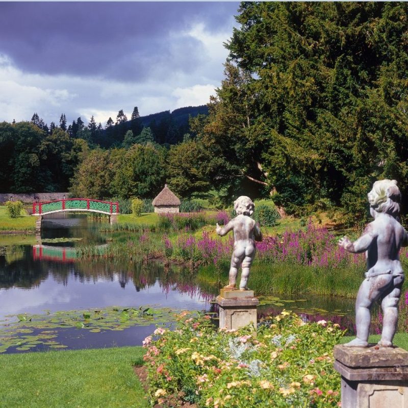Two stone cupid sculptures overlooking a large pond with a red and green decorative bridge, surrounded by bright flowers and lush green trees.