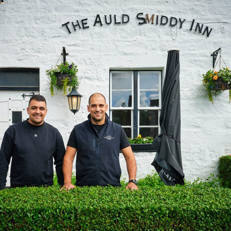 Two smiling men wearing all black standing outside a white building with the sign 'The Auld Smiddy Inn'.