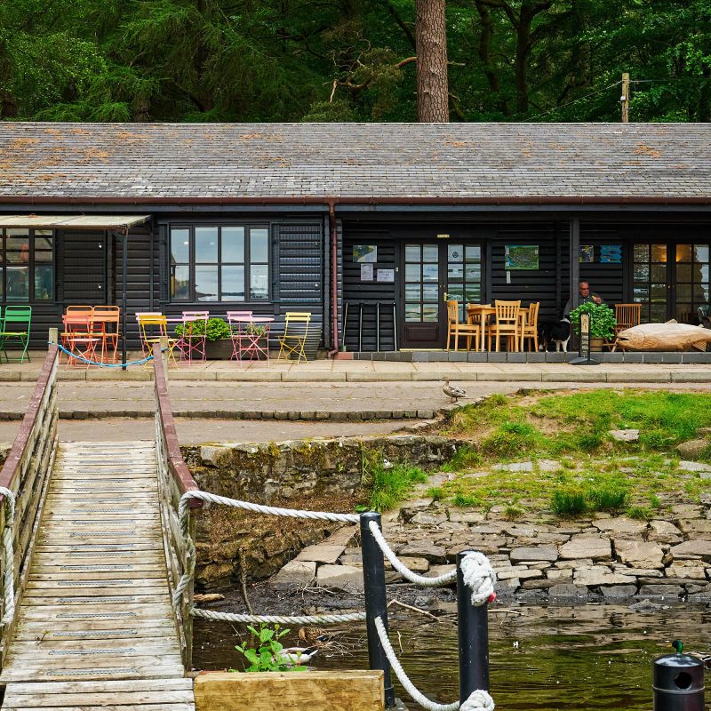 A black building with a small body of water in front of it and a wooden bridge.